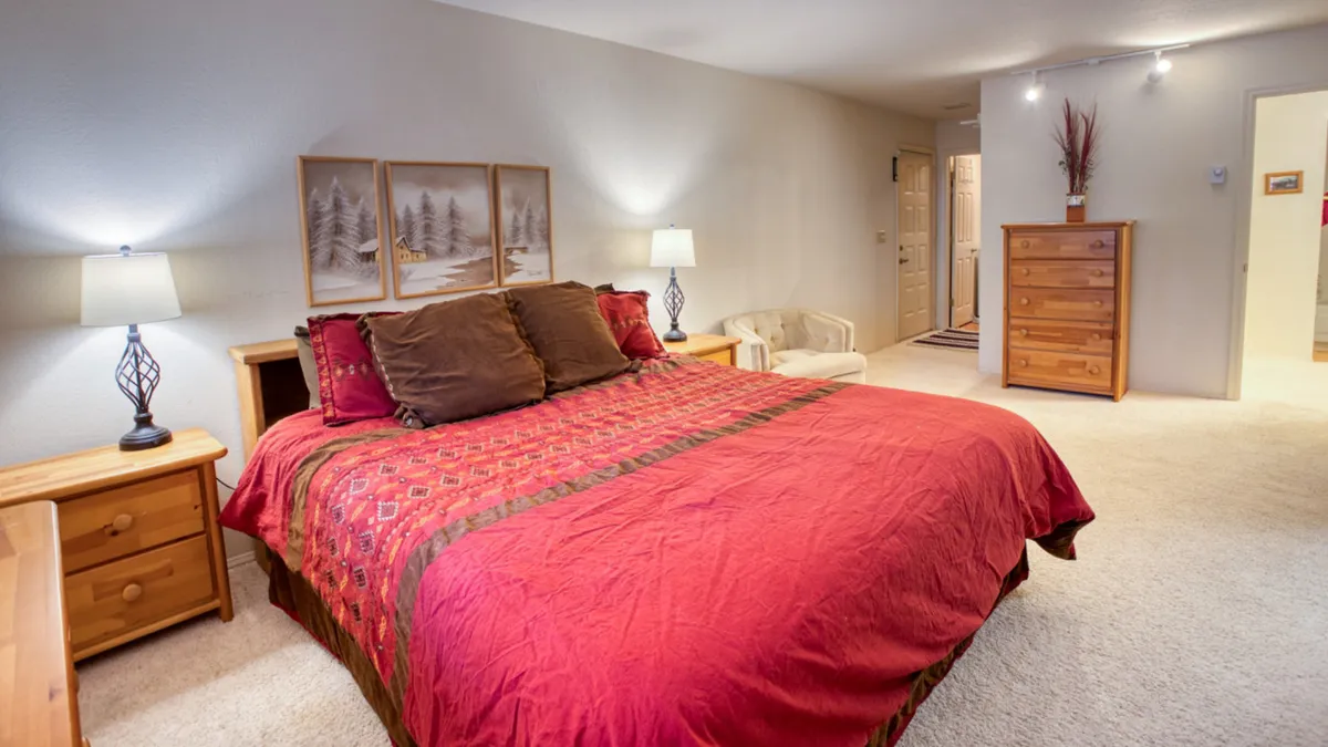 A bedroom with a full-size bed dressed in red and green bedding, flanked by nightstands with lamps and a wooden dresser. Decorative wall art adds a homely touch. The room feels inviting and warm, ideal for kids, friends, or grandparents.