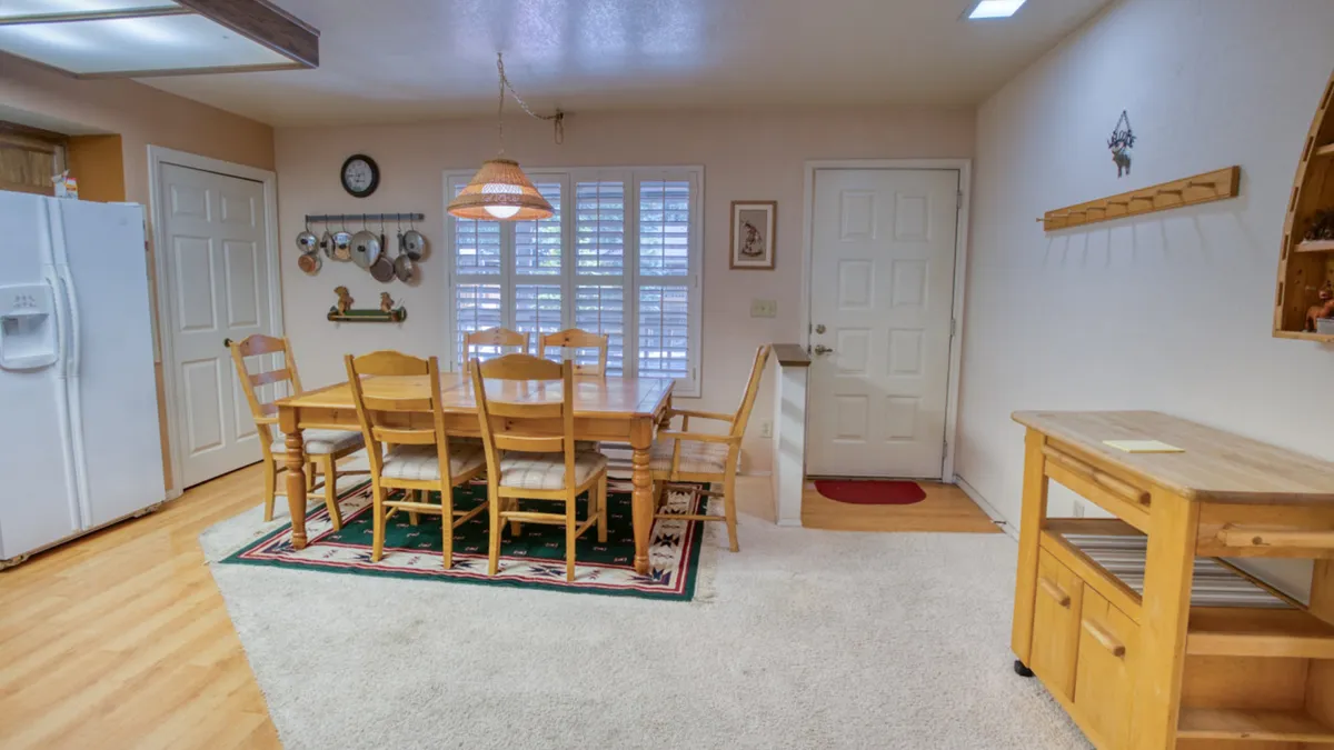 This image showcases a warm dining area with a wooden dining table, a hanging lamp casting a golden glow, kitchen utensils displayed on the wall, and a visible doorway leading to other home spaces. The setup invites family meals, celebrations, and shared moments around the table.