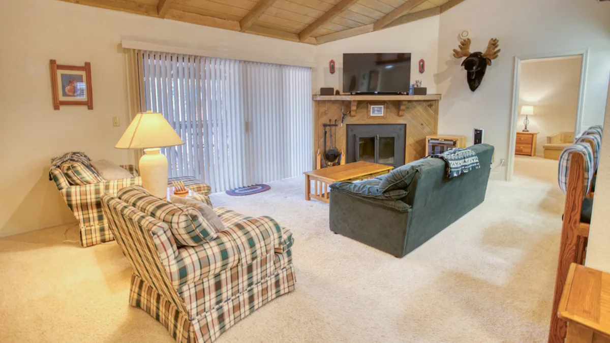 The image features a spacious living room with plaid sofas arranged around a wooden coffee table, a cozy fireplace, a television, and a decorative wall mount. The scene exudes warmth and comfort, inviting guests to relax, socialize, or enjoy evening entertainment together after a busy day outdoors.