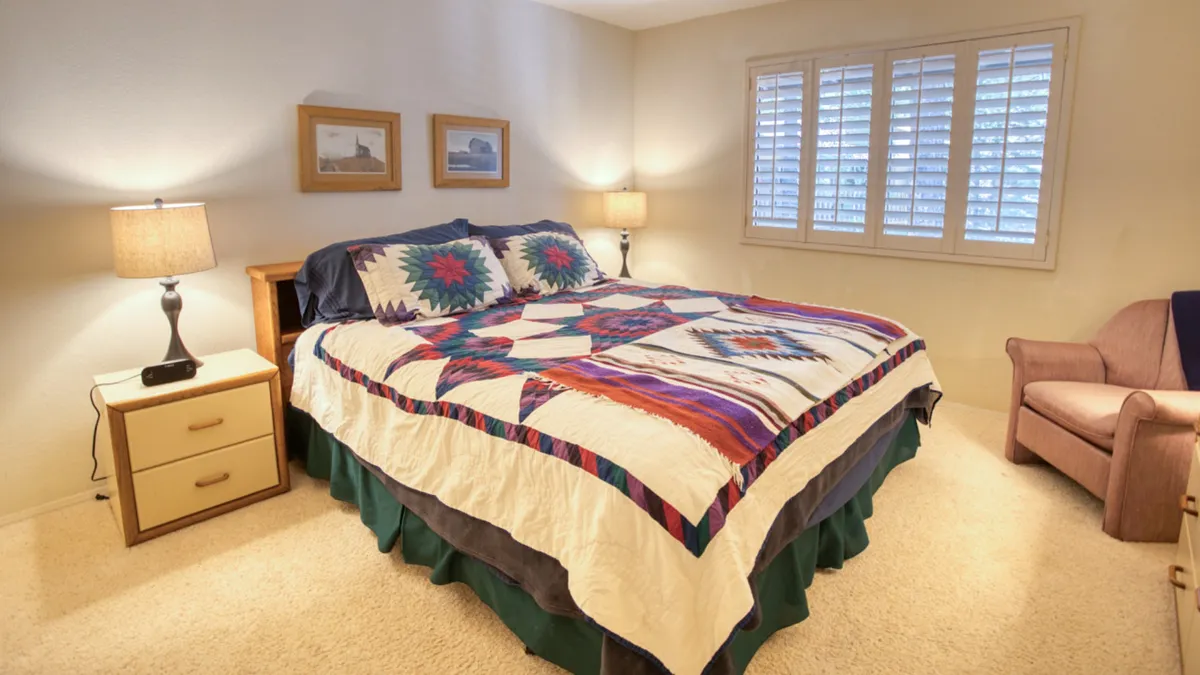 A cozy bedroom featuring a quilted bed bathed in natural light, flanked by nightstands with glowing lamps. An inviting armchair sits in the corner, and framed pictures add a personal touch to the warm, restful atmosphere.