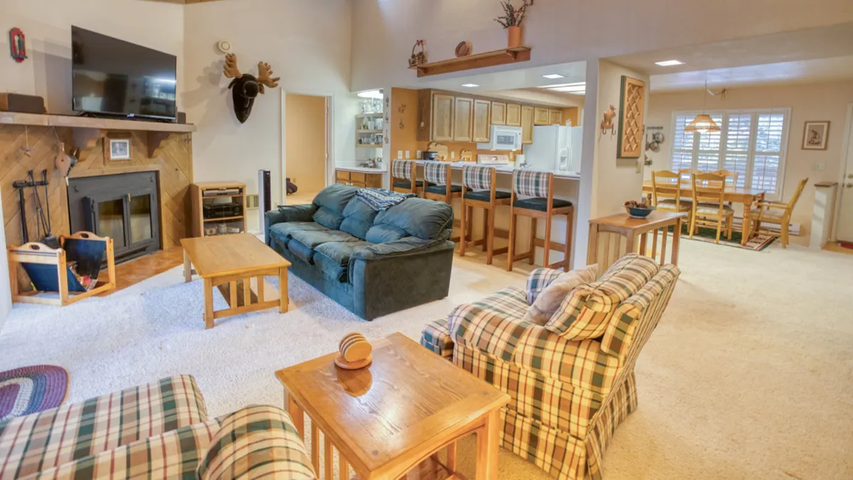 The image showcases a warm, inviting interior with a stone fireplace as the centerpiece, a plush sofa arranged for cozy gatherings, a dining area perfect for family meals, and a modern kitchen bar in view. The space suggests comfort, togetherness, and relaxation for families or groups.